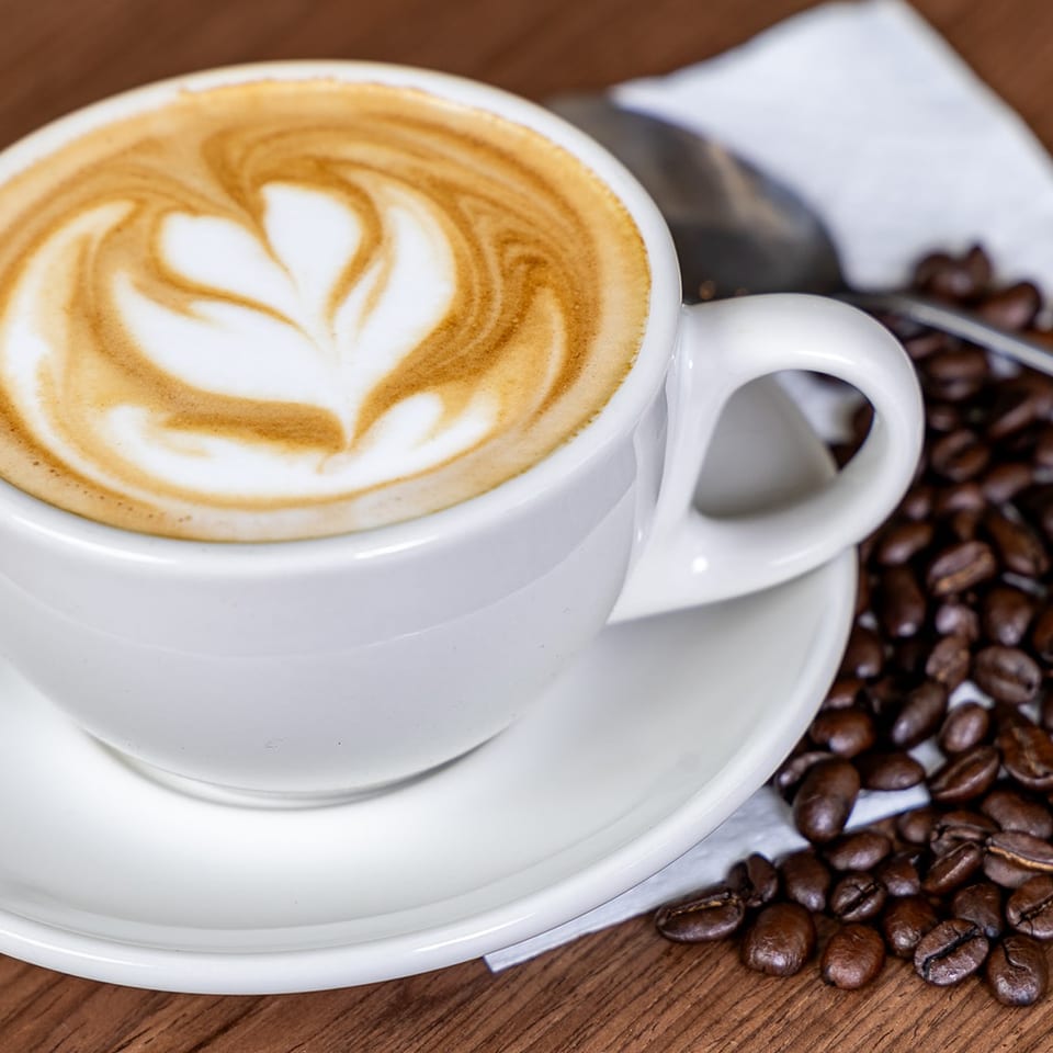 White cup of latte with leaf-shaped foam art on a saucer, surrounded by coffee beans and a spoon on a wooden table.