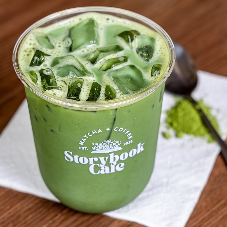 Iced green matcha drink in a Storybook Cafe cup, topped with ice, on a napkin beside a spoon and matcha powder.
