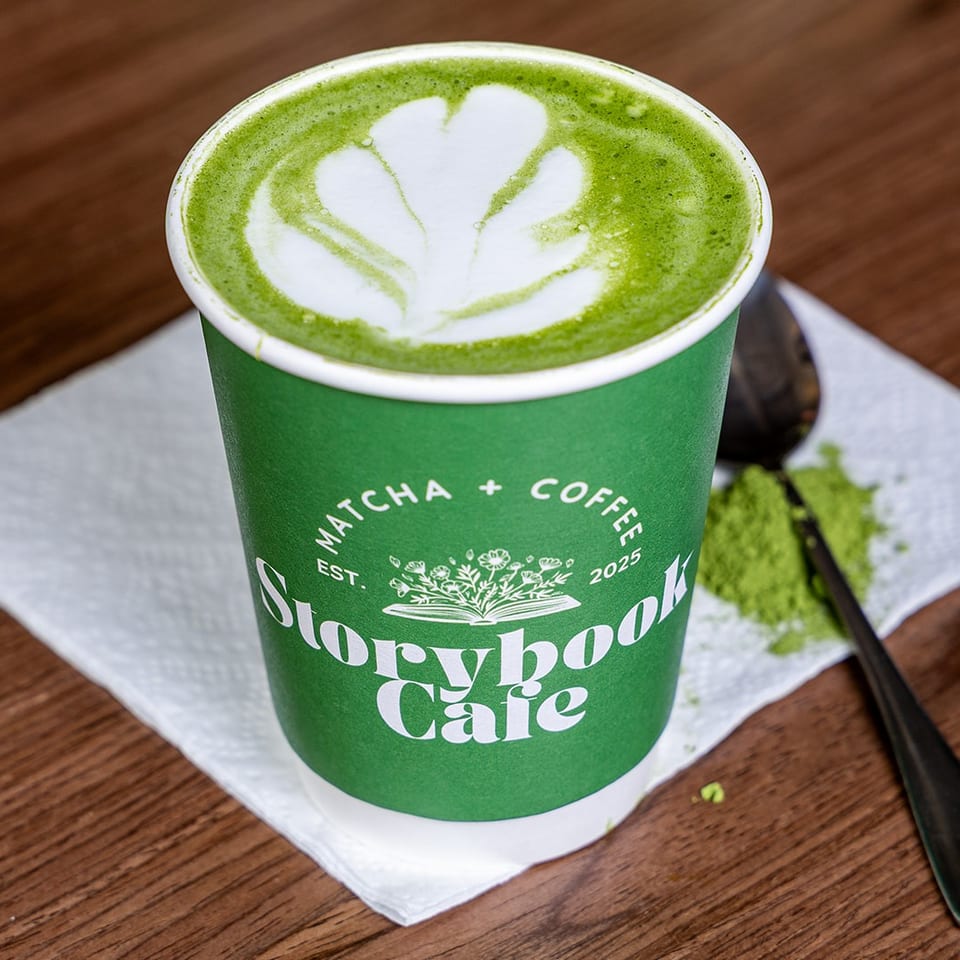 Green matcha latte with leaf-shaped foam art in a Storybook Cafe cup, beside a spoon and spilled matcha on a napkin.