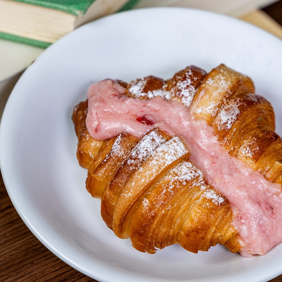 Golden croissant topped with pink berry cream and powdered sugar on a white plate beside books.