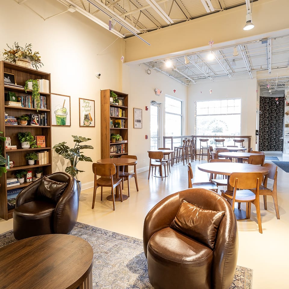 Bright, cozy café interior with wooden tables, leather lounge chairs, bookshelves, plants, artwork, and large windows.