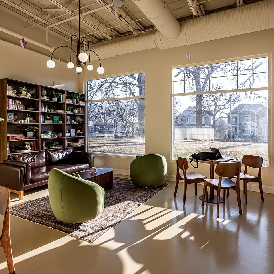 Bright restaurant lounge with leather sofa, green chairs, bookshelves, dining table, large windows, and exposed industrial ceiling.